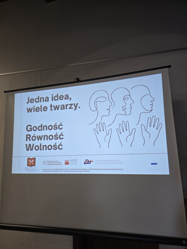 Na dużym ekranie wyświetlany jest biały slajd z tekstem i grafiką. U góry widnieje napis "Jedna idea, wiele twarzy.". Poniżej znajdują się słowa: "Godność, Równość, Wolność". Po prawej stronie widać trzy schematyczne rysunki profili głów i dłoni. Na dole slajdu widoczne są logotypy i mniejszy tekst.