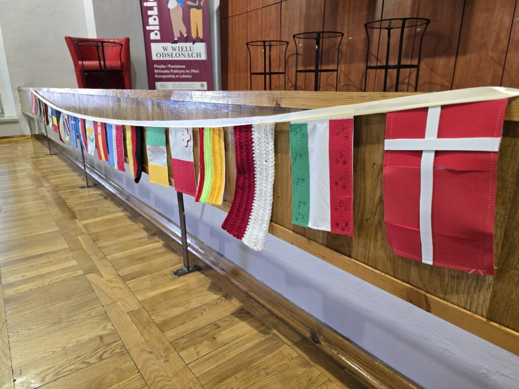 Długa szarfa z wiszącymi miniaturowymi flagami różnych krajów rozciągnięta wzdłuż drewnianej balustrady.