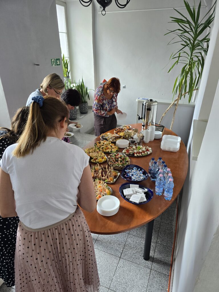 Pomieszczenie z jasnymi ścianami i wysokimi oknami. Na środku stoi owalny stół zastawiony różnymi potrawami, napojami i naczyniami. Kilka dorosłych osób obsługuje się przy stole.