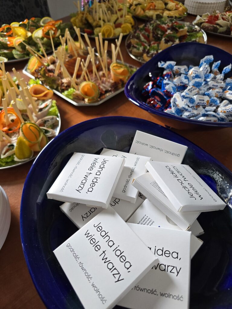 Stół zastawiony jest różnorodnymi przekąskami, takimi jak koreczki i kanapki. Na pierwszym planie, w dużej granatowej misie, znajduje się stos białych ulotek z napisem "Jedna idea, wiele twarzy. godność, równość, wolność". Obok widać misę z cukierkami w niebieskich papierkach.