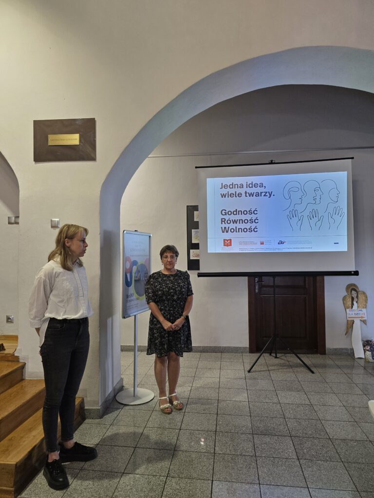 W pomieszczeniu z jasnymi ścianami i łukowatymi przejściami, na pierwszym planie stoi młoda blondynka. Dalej, pod dużym ekranem z prezentacją, stoi kobieta w ciemnej sukience. Na ekranie widoczny jest napis "Jedna idea, wiele twarzy. Godność Równość Wolność" oraz grafika trzech głów.