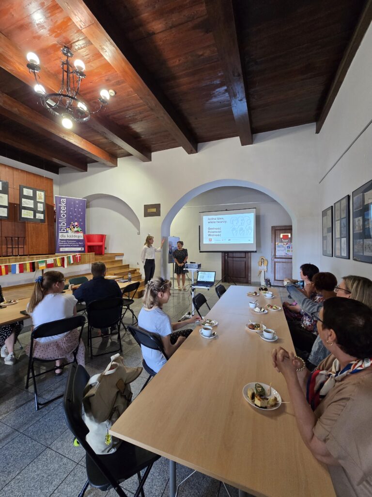 Duża sala z drewnianym sufitem i licznym audytorium. W tle, na podwyższeniu, dwie osoby prowadzą prezentację wyświetlaną na ekranie. Przed nimi siedzi wiele osób przy długich stołach, a na stołach widoczne są talerzyki z jedzeniem i napoje.