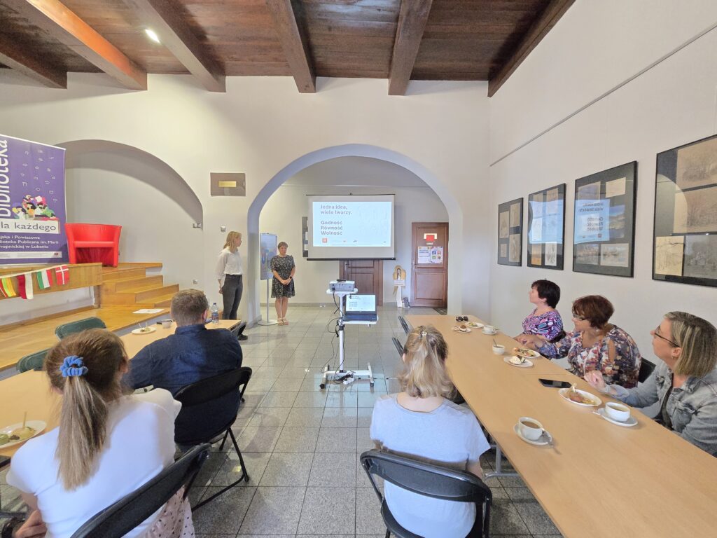 Duża sala z drewnianym sufitem i licznym audytorium. W głębi, pod ekranem z prezentacją, stoją dwie osoby. Przed nimi siedzi wiele osób przy długich stołach, na których znajdują się talerzyki z jedzeniem i napoje.