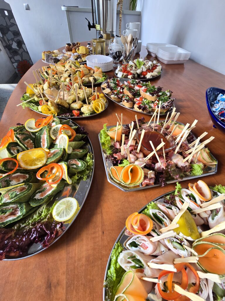 Drewniany stół zastawiony jest licznymi półmiskami z różnorodnymi przekąskami i przystawkami, w tym roladkami, koreczkami i tartinkami. W tle widoczne są naczynia i inne elementy cateringu.