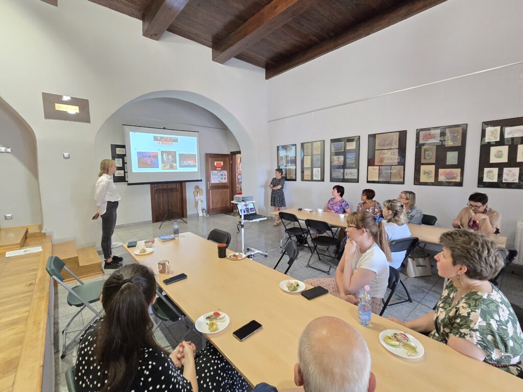 
Duża, jasna sala z drewnianym sufitem i ścianami ozdobionymi obrazami. W głębi sali, po lewej stronie, osoba stoi obok ekranu z wyświetlaną prezentacją.
