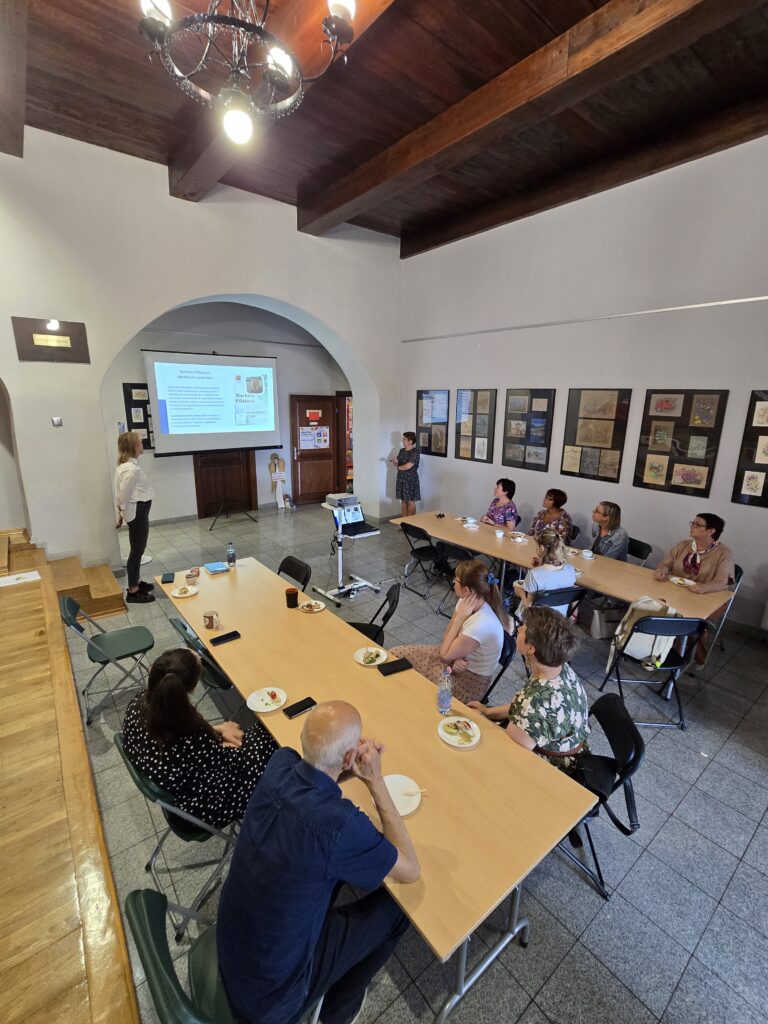 Duża sala z drewnianym sufitem i oknami. W głębi, pod ekranem z prezentacją, dwie osoby prowadzą wydarzenie. Przed nimi siedzi liczne audytorium przy długich stołach. Na stołach znajdują się talerzyki z jedzeniem i napoje. Ściany są ozdobione obrazami.