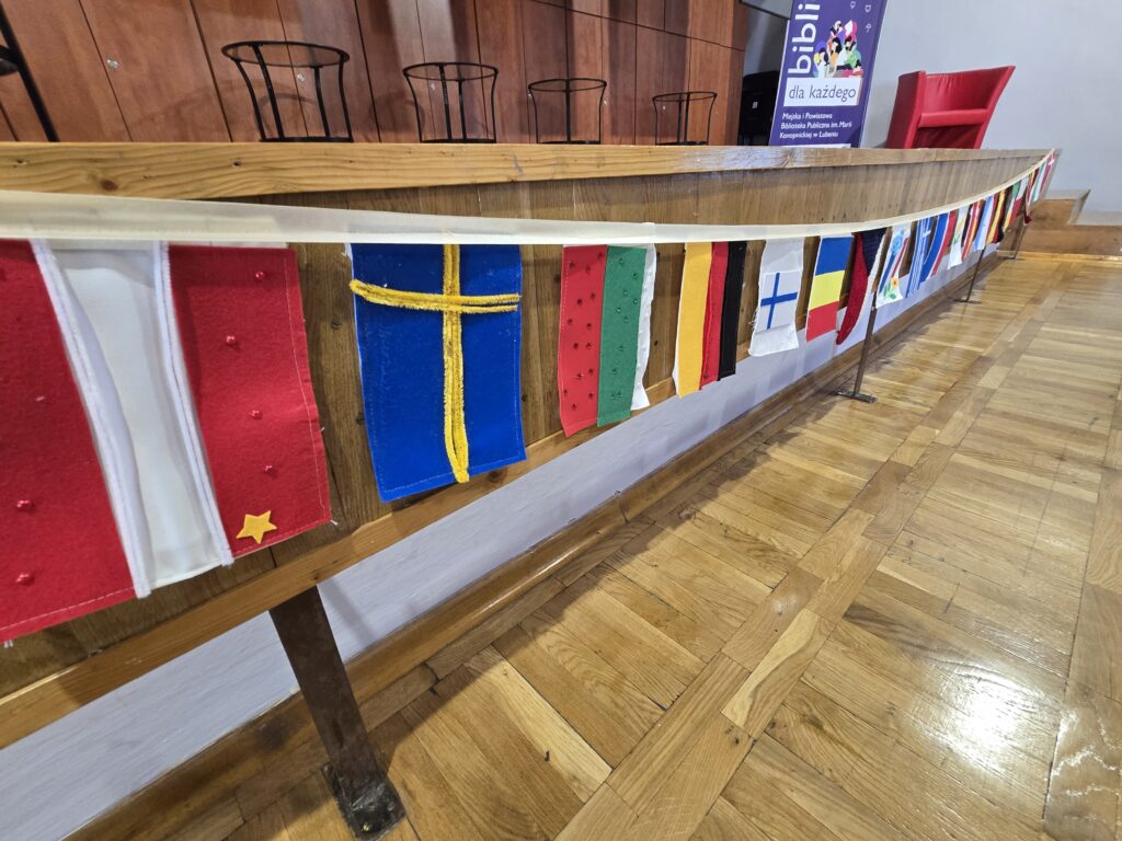 Szereg małych, kolorowych flag różnych krajów jest zawieszony na drewnianej balustradzie w pomieszczeniu z drewnianą podłogą.
