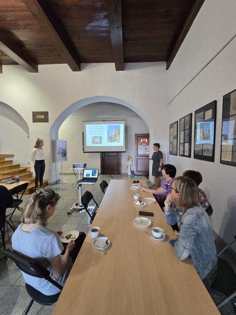 Duża sala z drewnianym sufitem i łukowatymi przejściami. W głębi, pod ekranem z prezentacją, stoją dwie kobiety. Przed nimi siedzi wiele osób przy długich stołach. 