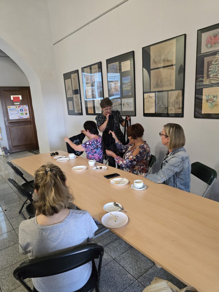 Pięć kobiet siedzi przy długim, drewnianym stole w jasnym pomieszczeniu. Dwie kobiety w środku oglądają coś w torebce. 