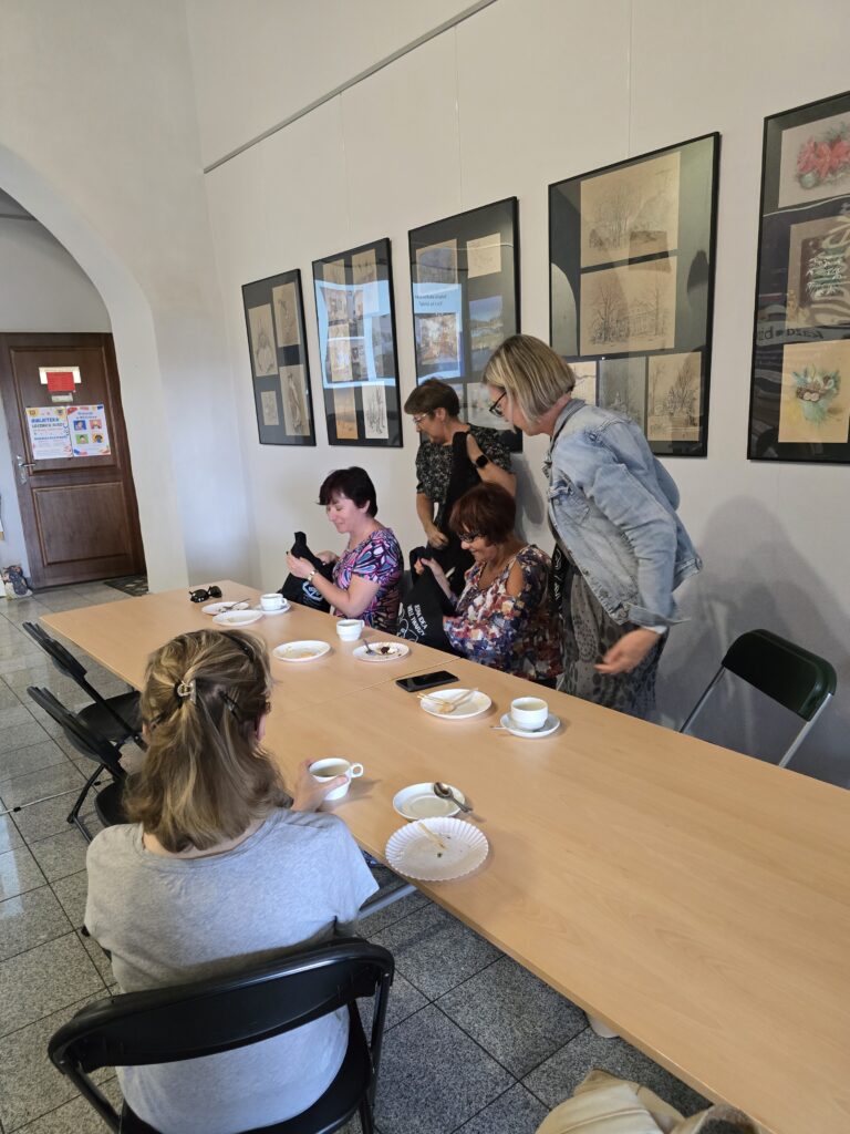 Pięć kobiet siedzi lub stoi przy długim, drewnianym stole w jasnym pomieszczeniu. Dwie kobiety w środku oglądają coś w torebkach, a inna kobieta w dżinsowej kurtce stoi obok.