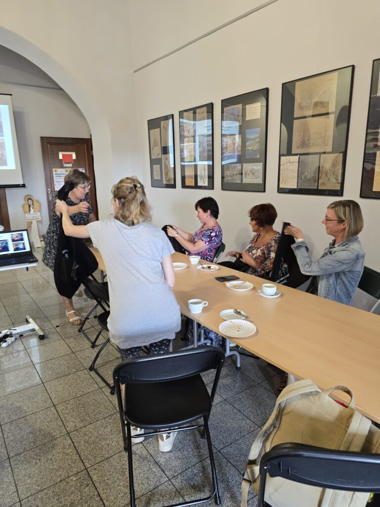 Pięć kobiet siedzi lub stoi przy długim, drewnianym stole w jasnym pomieszczeniu. Dwie kobiety w centrum zajmują się oglądaniem zawartości toreb.