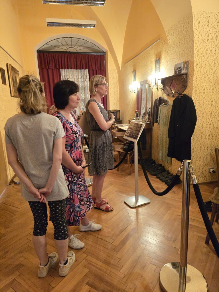 Trzy kobiety stoją na drewnianej podłodze w pokoju z żółtymi ścianami i łukowatym sufitem. Oglądają wystawę, na której widać ubrania, lustra i inne eksponaty.