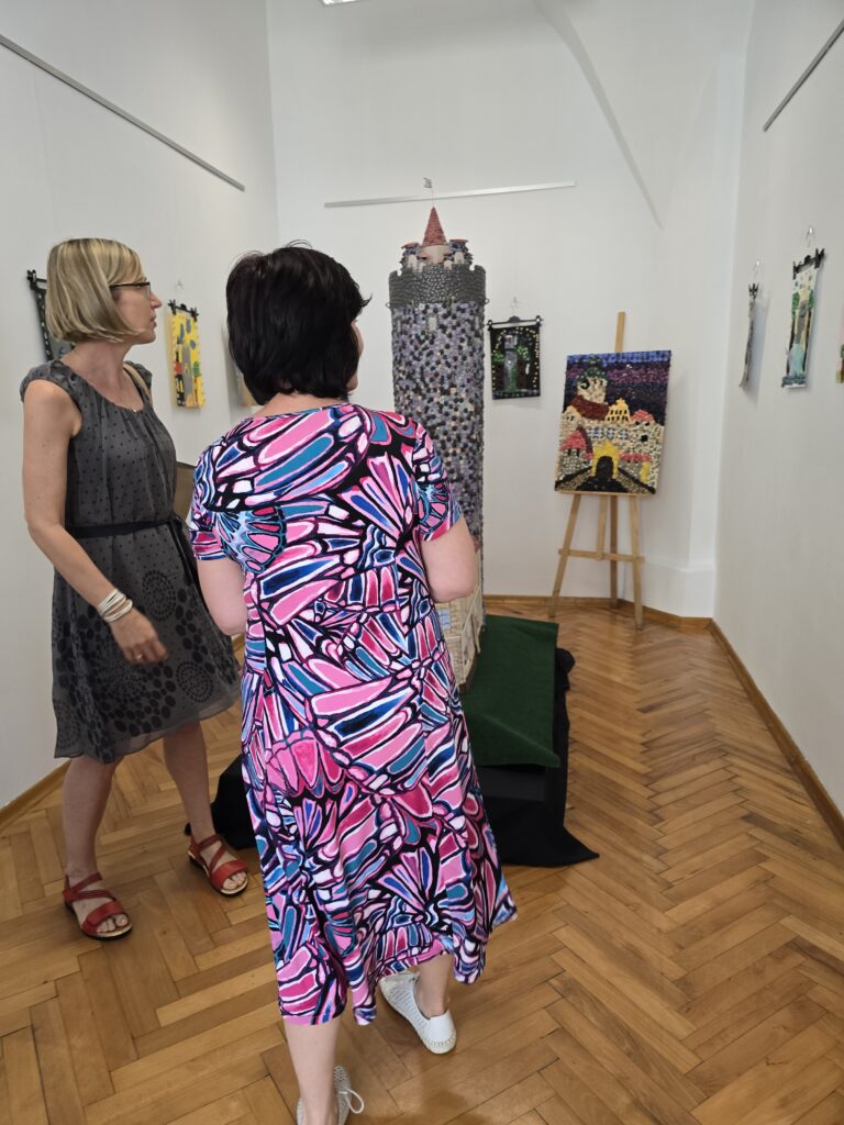 Dwie kobiety oglądają wystawę sztuki w jasnym pomieszczeniu z drewnianą podłogą. Jedna kobieta w szarej sukience patrzy w lewo, druga, w kolorowej sukience, stoi tyłem do obiektywu, patrząc na centralnie umieszczoną, wysoką rzeźbę. W tle widoczne są również obrazy na ścianach i sztaluga z dziełem.