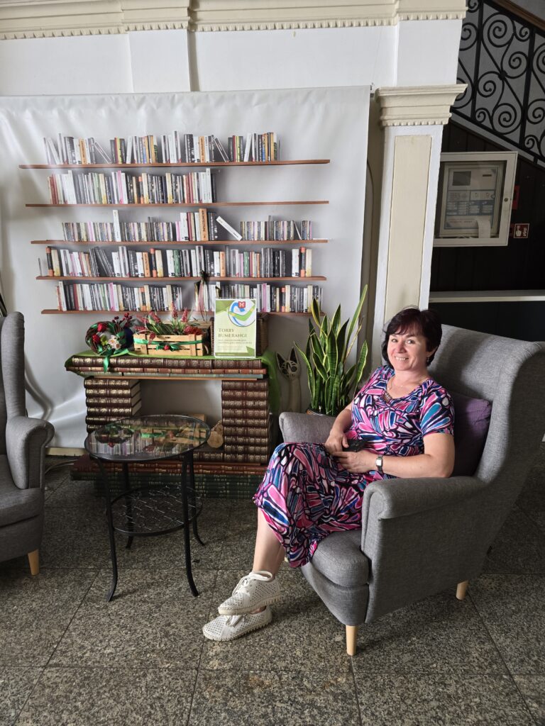 Kobieta w kolorowej sukience siedzi w szarym fotelu, uśmiechając się. Za nią znajduje się duży plakat przedstawiający regały z książkami. Obok fotela stoi stolik z doniczką i dekoracjami.