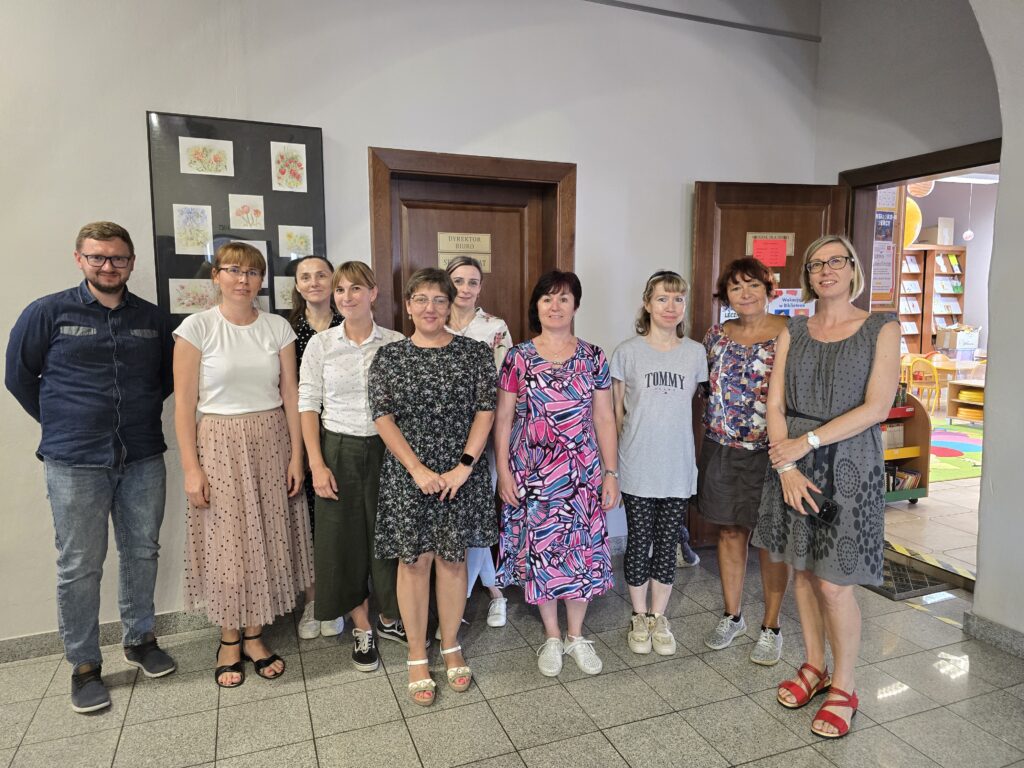 Grupa dziesięciu dorosłych osób – jeden mężczyzna i dziewięć kobiet – stoi razem w holu przed brązowymi drzwiami. 