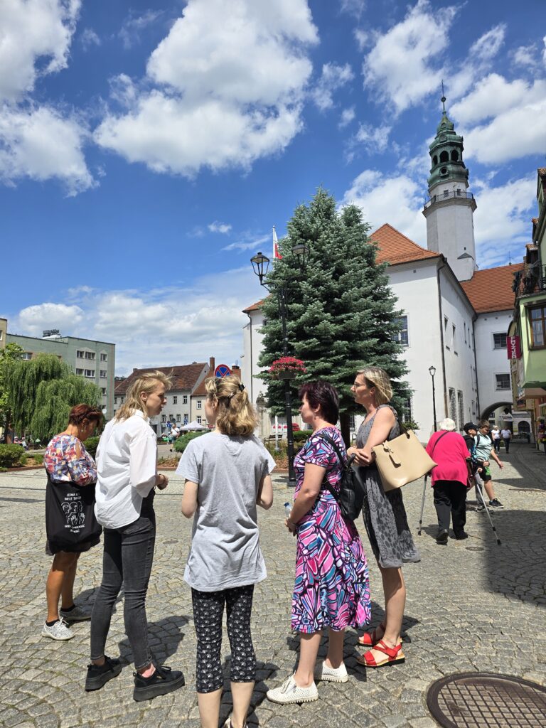 Grupa pięciu kobiet rozmawia na brukowanym placu w słoneczny dzień. W tle widoczny jest wysoki budynek z zielonym dachem, duże drzewo iglaste oraz budynki miejskie.