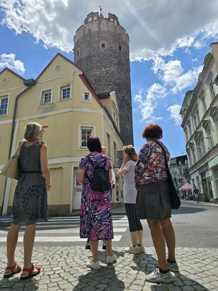 Cztery kobiety stoją na brukowanym placu z przejściem dla pieszych, patrząc w górę na wysoką kamienną wieżę. Po lewej stronie widać żółty budynek. Niebo jest niebieskie z białymi chmurami.