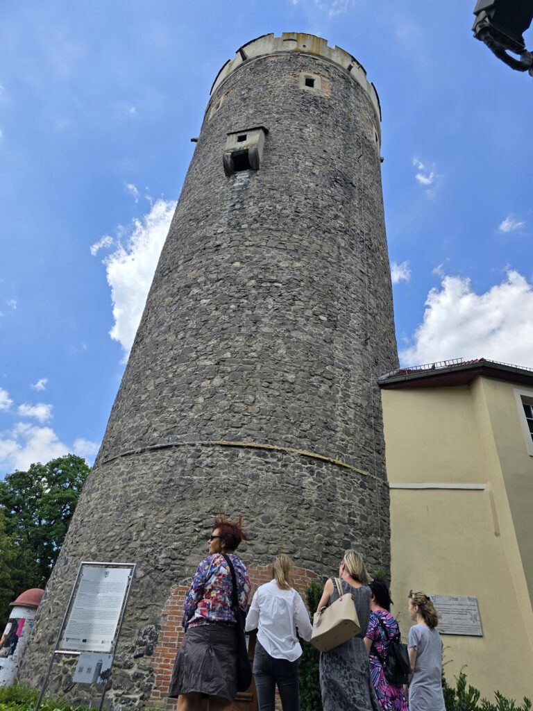 Grupa czterech kobiet stoi u podnóża wysokiej, kamiennej wieży, patrząc w górę. Obok wieży widoczny jest fragment jasnego budynku. Niebo jest błękitne z białymi chmurami.