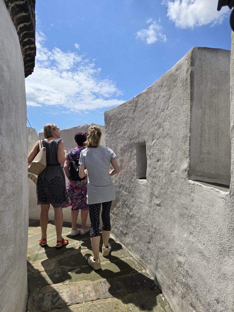 Trzy kobiety idą wąskim, brukowanym korytarzem między wysokimi, jasnymi murami. Słońce rzuca długie cienie na ziemię. W górze widać fragment błękitnego nieba z chmurami.