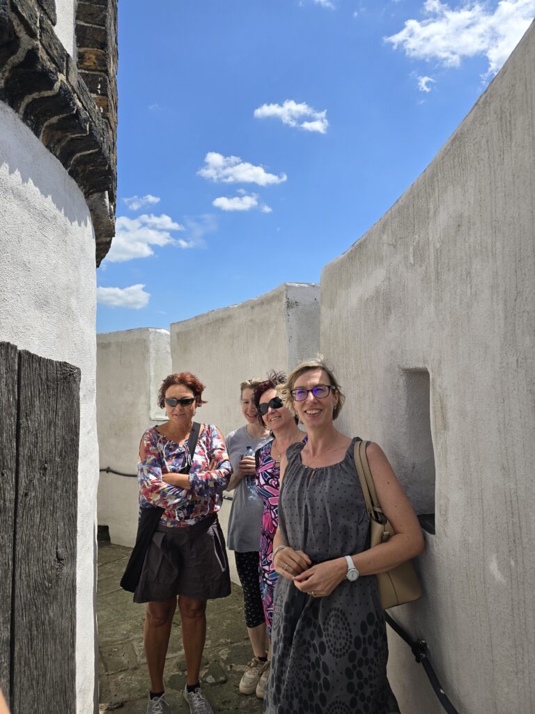 Cztery kobiety uśmiechają się do obiektywu, stojąc w wąskim przejściu między jasnymi murami. Niebo jest błękitne z białymi chmurami.