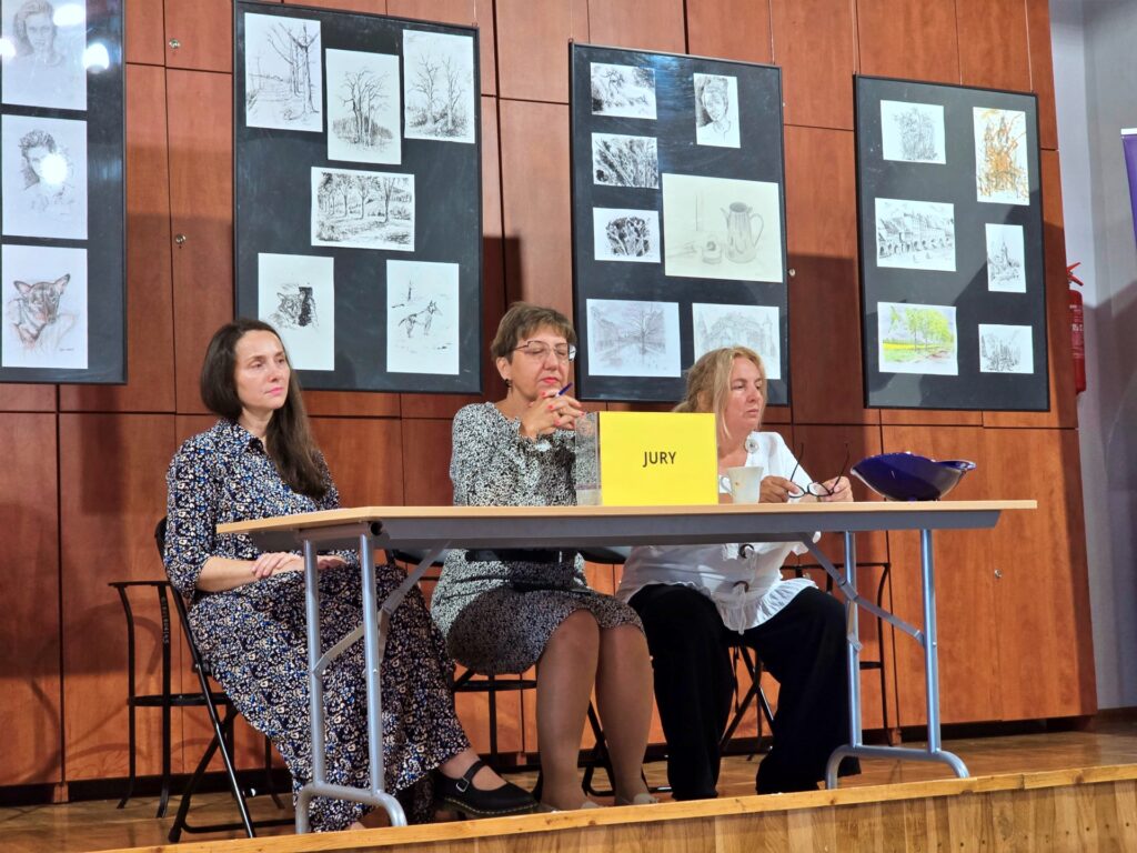 Trzy dorosłe kobiety siedzą za stołem na podwyższeniu, na którym stoi żółta tabliczka z napisem "JURY". W tle widoczna jest drewniana ściana z wieloma oprawionymi grafikami i rysunkami.
