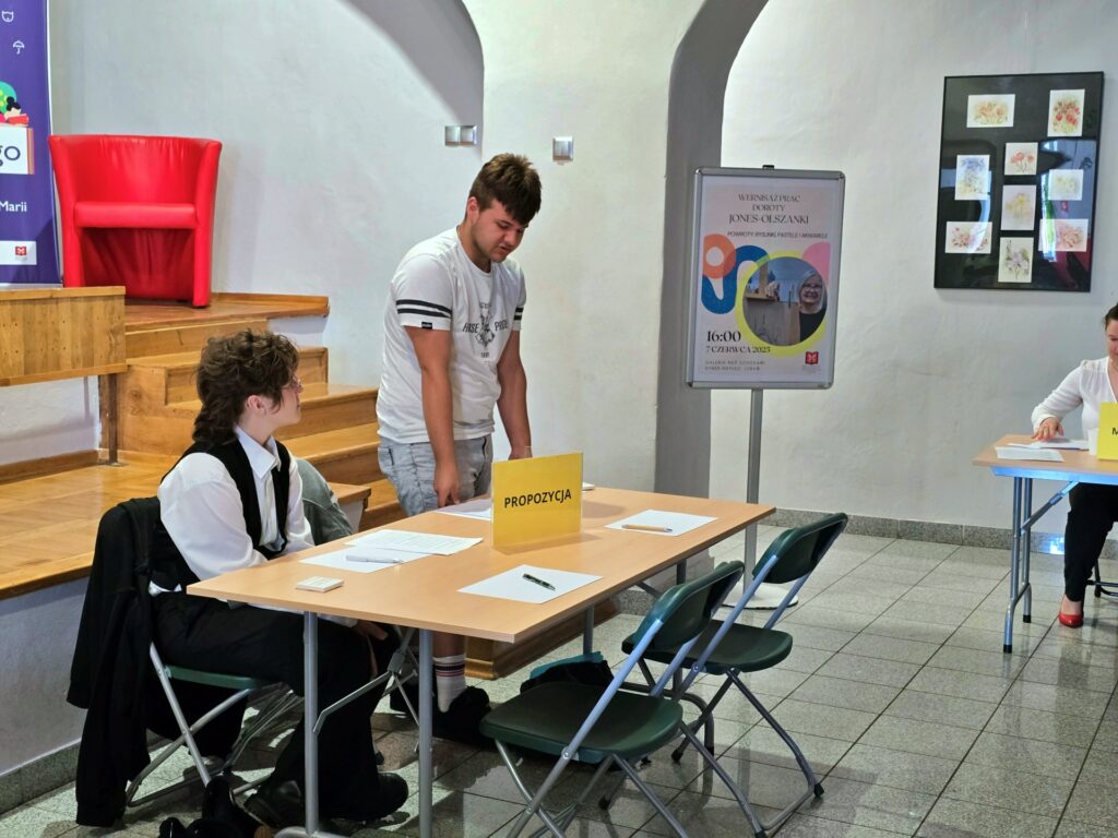 Wnętrze jasnego pomieszczenia z drewnianymi schodami i czerwonym fotelem po lewej stronie. Przy stole z tabliczką "PROPOZYCJA" siedzi młoda osoba, a obok niej stoi inny młody mężczyzna. Po prawej stronie widoczna jest tablica informacyjna z plakatem.