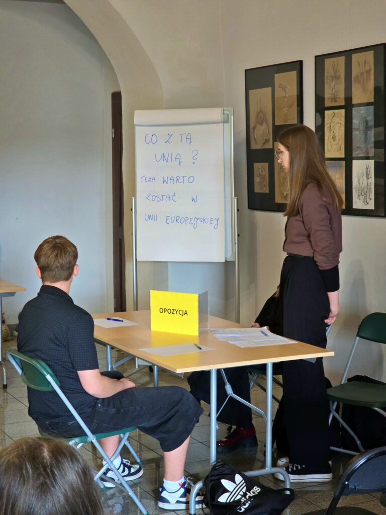 W sali ze ścianą ozdobioną obrazami, młoda kobieta stoi obok stołu z tabliczką "OPOZYCJA". Obok niej siedzi młody mężczyzna, a w tle widać białą tablicę z napisem.