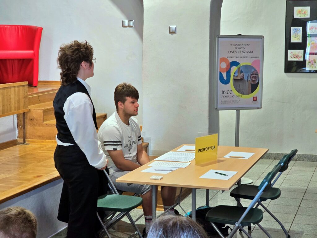 Jasnoszare pomieszczenie z drewnianymi schodami po lewej stronie, na których stoi czerwony fotel. Młoda osoba w czarno-białej kamizelce stoi obok stołu. Przy stole siedzi młody mężczyzna w jasnej koszulce. Na stole leży tabliczka z napisem "PROPOZYCJA". Po prawej stronie widoczna jest tablica informacyjna z plakatem.