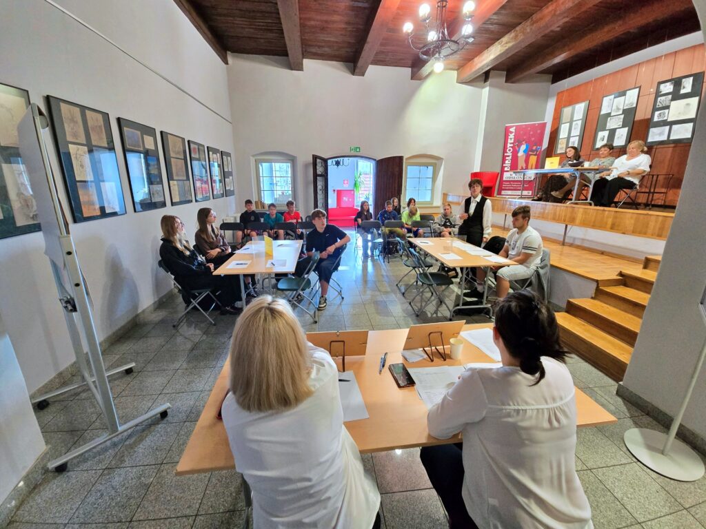 Duża sala z drewnianym sufitem i wieloma osobami. Na pierwszym planie widoczne są plecy dwóch osób siedzących przy stole. W głębi, przy kilku stołach, siedzi grupa ludzi, a na podwyższeniu po prawej stronie siedzi kilka osób. Z tyłu widoczny jest regał z napisem "BIBLIOTEKA".