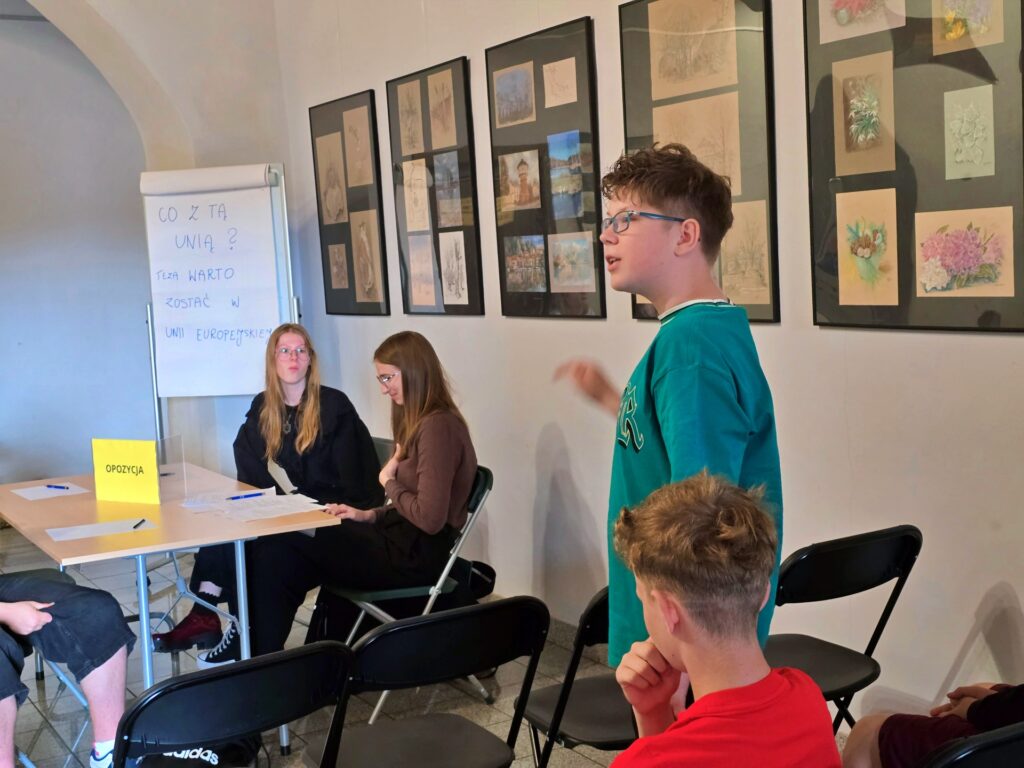 Na pierwszym planie, po prawej stronie, stoi młody chłopak w okularach i turkusowej bluzie, gestykulując. Po lewej stronie, za stołem z napisem "OPOZYCJA", siedzą dwie młode kobiety. W tle widoczna jest biała tablica z tekstem oraz ściana z oprawionymi grafikami.