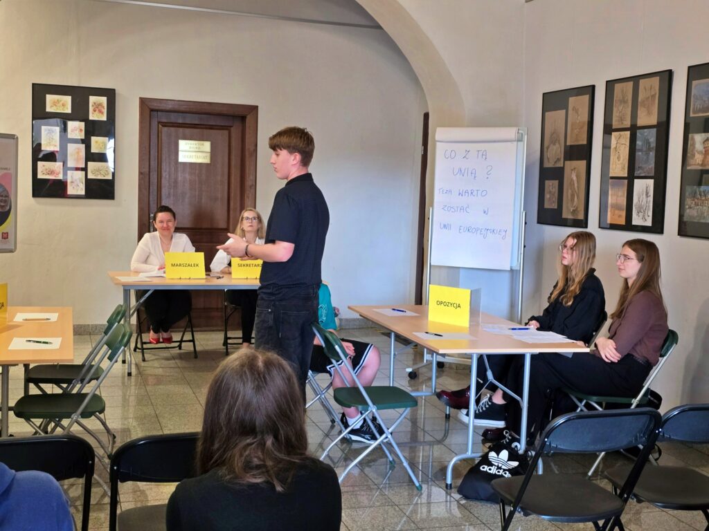 Duża sala z beżowymi ścianami i brązowymi drzwiami. Po lewej stronie, za stołem, siedzą dwie kobiety. Jedna z nich ma tabliczkę z napisem "MARSZAŁEK", a druga "SEKRETARZ". Na środku zdjęcia stoi młody mężczyzna w czarnej koszulce i dżinsach, zwrócony w stronę widowni. Po prawej stronie, za stołem, siedzą trzy młode kobiety. Nad stołem widoczna jest tablica z pytaniami. Na ścianach wiszą oprawione obrazy.