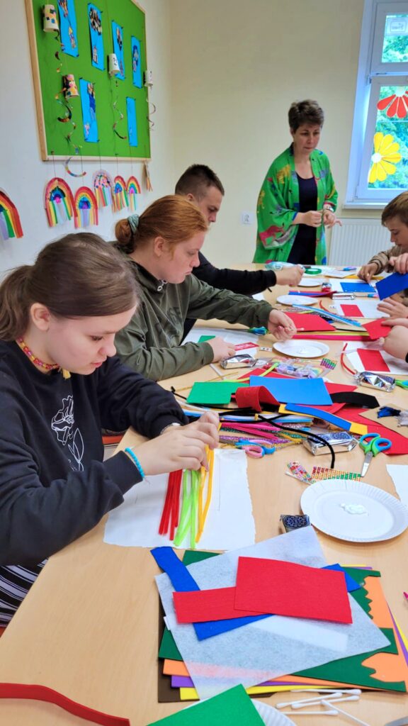 W sali z zieloną tablicą, przy drewnianym stole siedzi troje dzieci, każde zajęte tworzeniem rękodzieła z kolorowego papieru i innych materiałów. Dwie dorosłe osoby nadzorują ich pracę. Na stole leżą nożyczki, klej, bibuła, filc i gotowe prace.