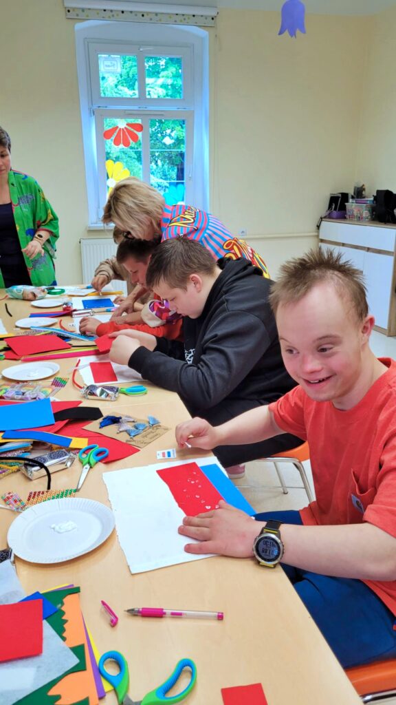 W jasnej sali, przy drewnianym stole siedzi grupa młodych ludzi i dorosłych, zajętych tworzeniem prac plastycznych z kolorowego papieru i bibuły. Na stole leżą nożyczki, klej, wycięte kształty i inne materiały. Osoba na pierwszym planie po prawej stronie uśmiecha się, patrząc na swoją pracę.