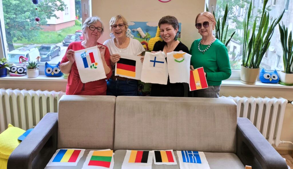 Cztery uśmiechnięte kobiety trzymają własnoręcznie wykonane, materiałowe flagi różnych państw. Na kanapie przed nimi leżą kolejne, mniejsze flagi.