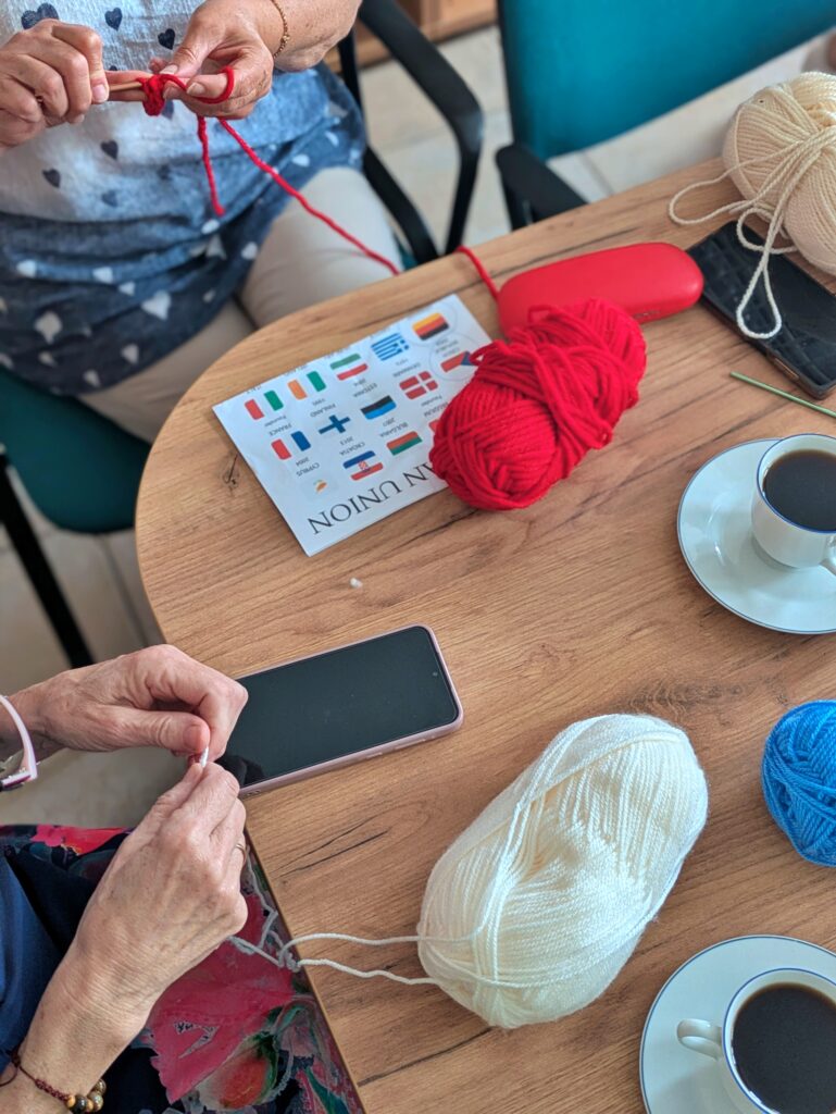 Zbliżenie na drewniany stół, na którym leżą dwie szpule włóczki w kolorach czerwonym i kremowym. Pośrodku stołu leży biała kartka z wydrukowanymi flagami państw, a obok niej czerwone etui na okulary i telefon komórkowy. Widać ręce dwóch osób robiących na drutach. Obok leżą dwie białe filiżanki z kawą.