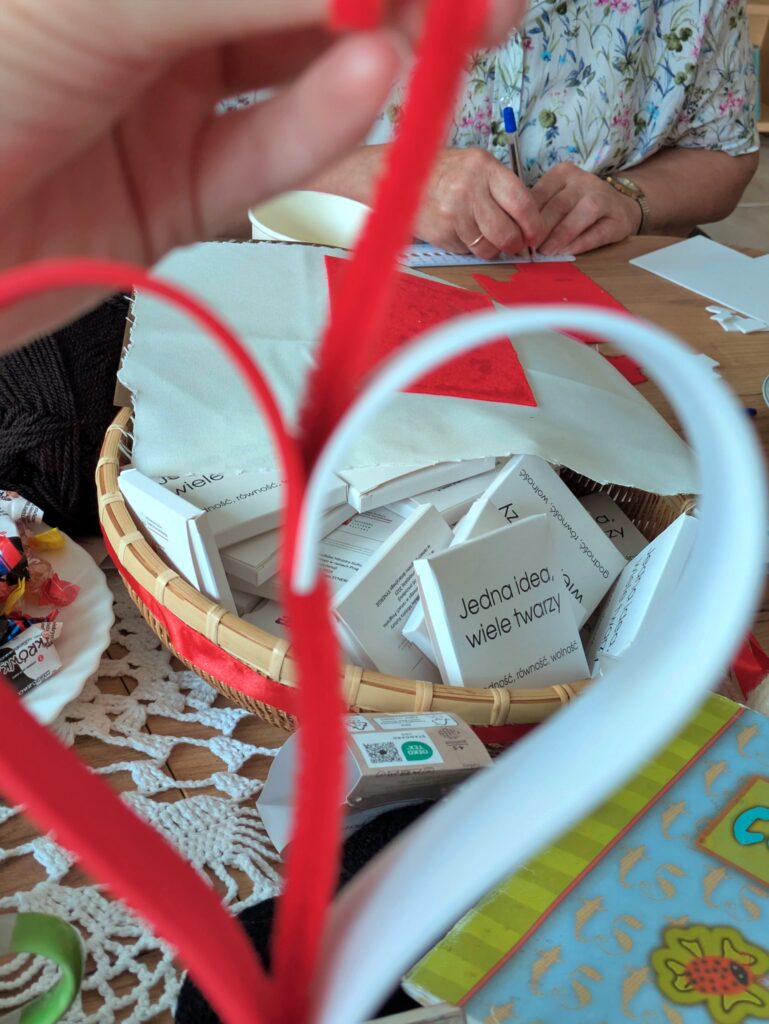 Zbliżenie na ręce trzymające dwa kształty serca wykonane z czerwonej i białej wstążki. W tle widać wiklinowy koszyk wypełniony małymi, białymi ulotkami z napisem "Jedna idea, wiele twarzy". Obok leżą słodycze w papierkach. Po prawej stronie widać fragment otwartego zeszytu.