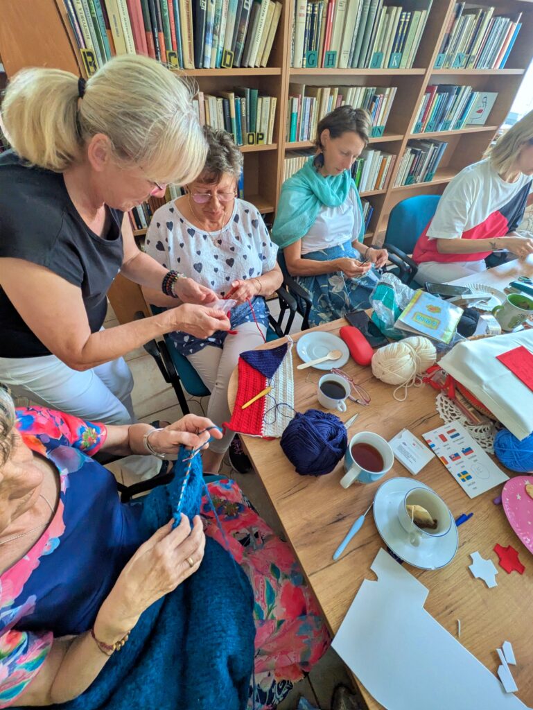 Sześć kobiet siedzi przy stole, zajętych robótkami ręcznymi. Na stole leży włóczka, druty i szydełko.