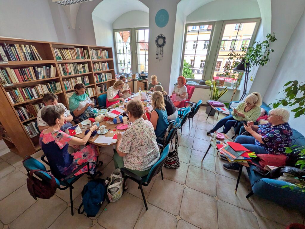 Na zdjęciu widać grupę około dziesięciu kobiet, które siedzą w pomieszczeniu wypełnionym regałami z książkami, zajmując się różnymi formami rękodzieła.