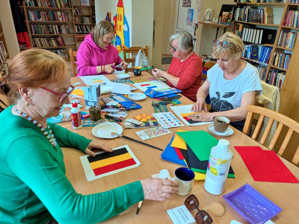 Cztery kobiety siedzą przy stole, zajęte pracami ręcznymi. Przed nimi rozłożone są różne materiały, w tym kolorowe tkaniny, kleje i przybory do tworzenia.