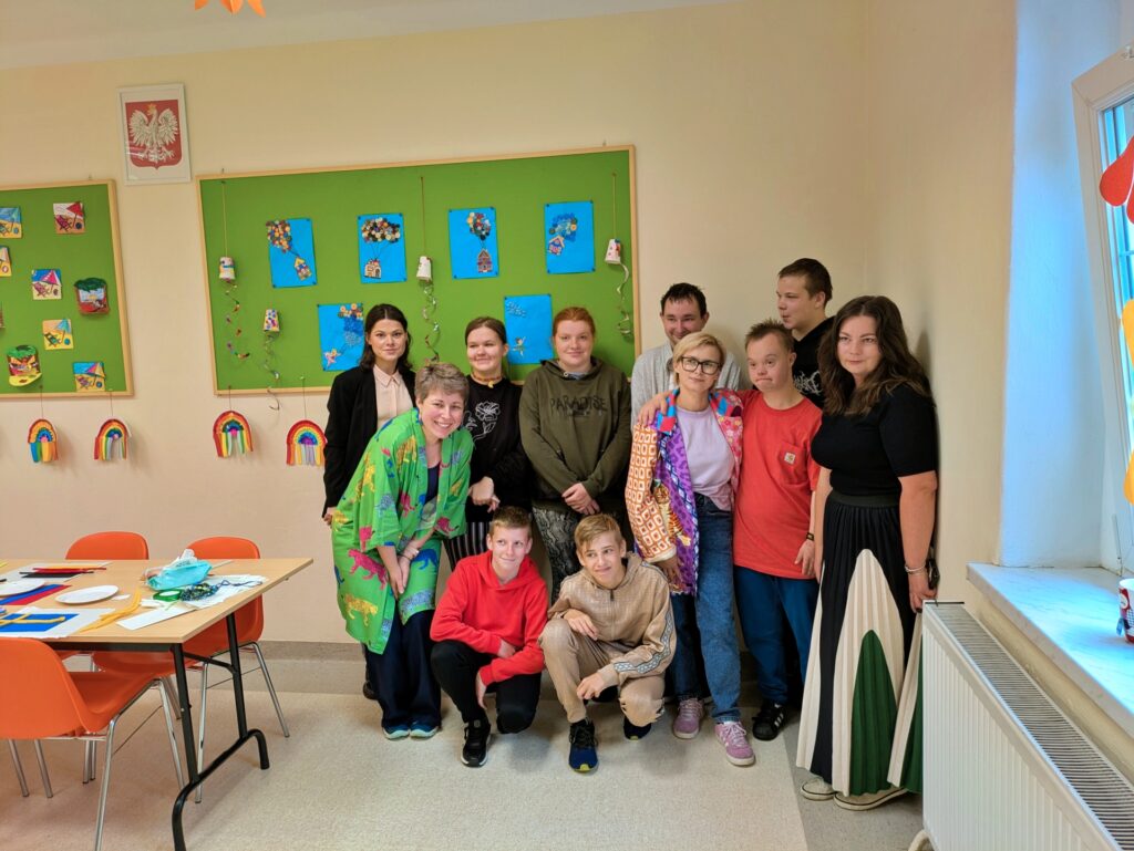 Na zdjęciu widać grupę dwunastu osób, stojących i kucających. Po lewej stronie zdjęcia widać pomarańczowe stoły i krzesła.