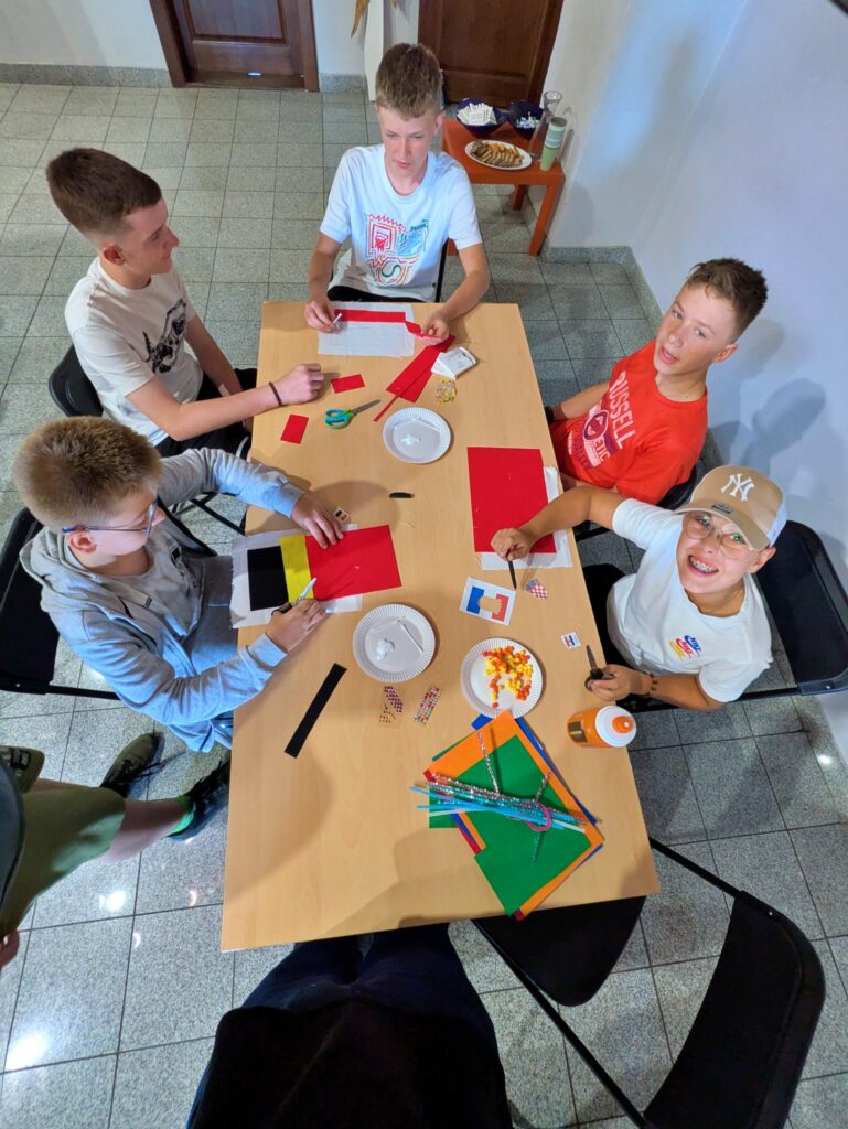 Pięciu młodych chłopców siedzi wokół drewnianego stołu, zajętych tworzeniem rękodzieła z papieru. Na stole leżą kolorowe arkusze papieru, nożyczki, klej i różne ozdoby.