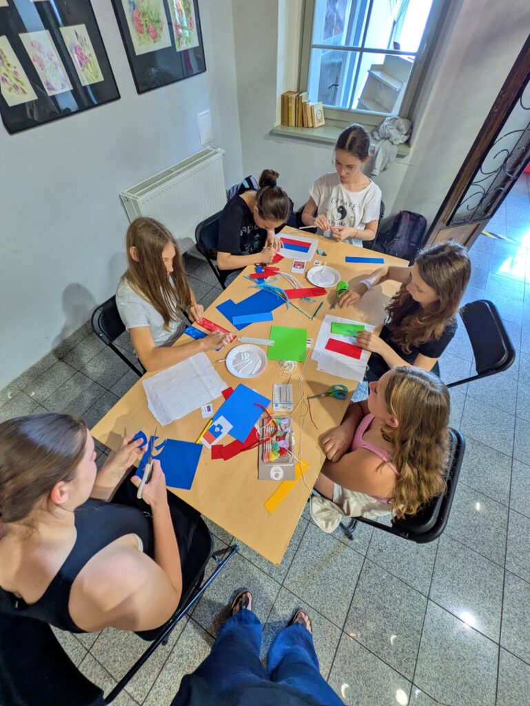 Sześć młodych dziewcząt siedzi przy stole, zajętych tworzeniem rękodzieła. Na stole leżą kolorowe arkusze papieru, nożyczki, klej i inne akcesoria plastyczne.